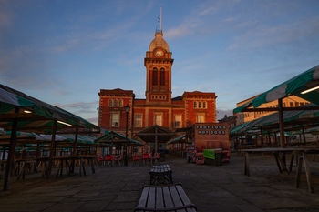 Chesterfield market hall morning The image shows Chesterfield Market Hall in Derbyshire, United Kingdom during a winter morning, with the sun casting a warm light on the architecture of the historic building. The urban setting is characterised by empty market stalls covered with green and white awnings, and the market hall's prominent clock tower rising above the square. Trademark signage and benches are visible in the foreground, highlighting the commercial function of this central location. This urban architecture photograph captures the quiet atmosphere of Chesterfield Market Hall before the market becomes busy, emphasising its role as a significant landmark in Derbyshire.